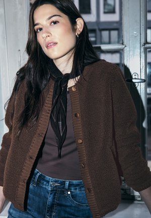 Giovane donna con lunghi capelli scuri che indossa una giacca in pile marrone con bottoni, una sciarpa scura e jeans blu, in piedi all'interno vicino a una finestra.