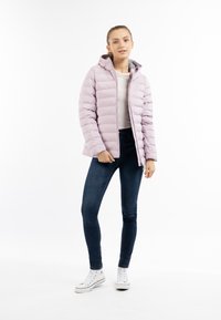 Giacca a vento di colore lilla chiaro con cuciture orizzontali, cappuccio e zip frontale. Indossata sopra una maglietta bianca e jeans scuri con sneakers bianche.