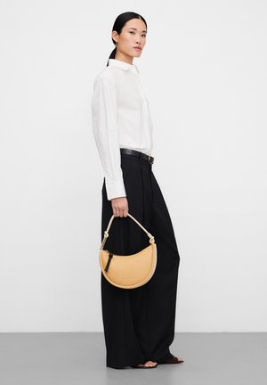 Donna con capelli neri, indossa una camicia bianca e pantaloni neri larghi, tiene una borsa a mezza luna beige con una tracolla corta.