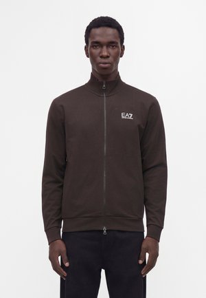 Homme portant une veste zippée marron foncé avec le logo EA7 Emporio Armani sur la poitrine gauche, debout devant un fond blanc uni.