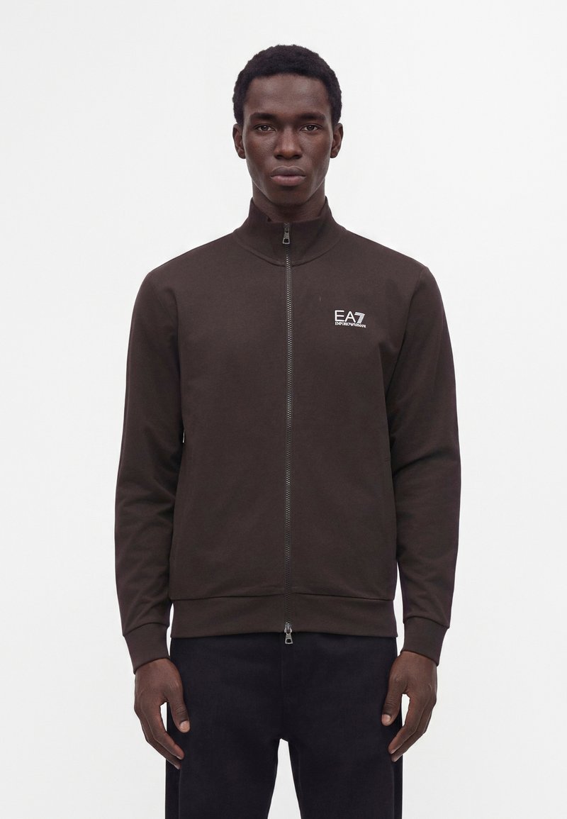 Homme portant une veste zippée marron foncé avec le logo EA7 Emporio Armani sur la poitrine gauche, debout devant un fond blanc uni.
