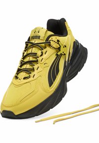 Baskets Puma jaunes et noires avec lacets noirs, breloque Pikachu sur le côté, visage de Pikachu sur la languette et lacets jaunes supplémentaires à côté de la chaussure.