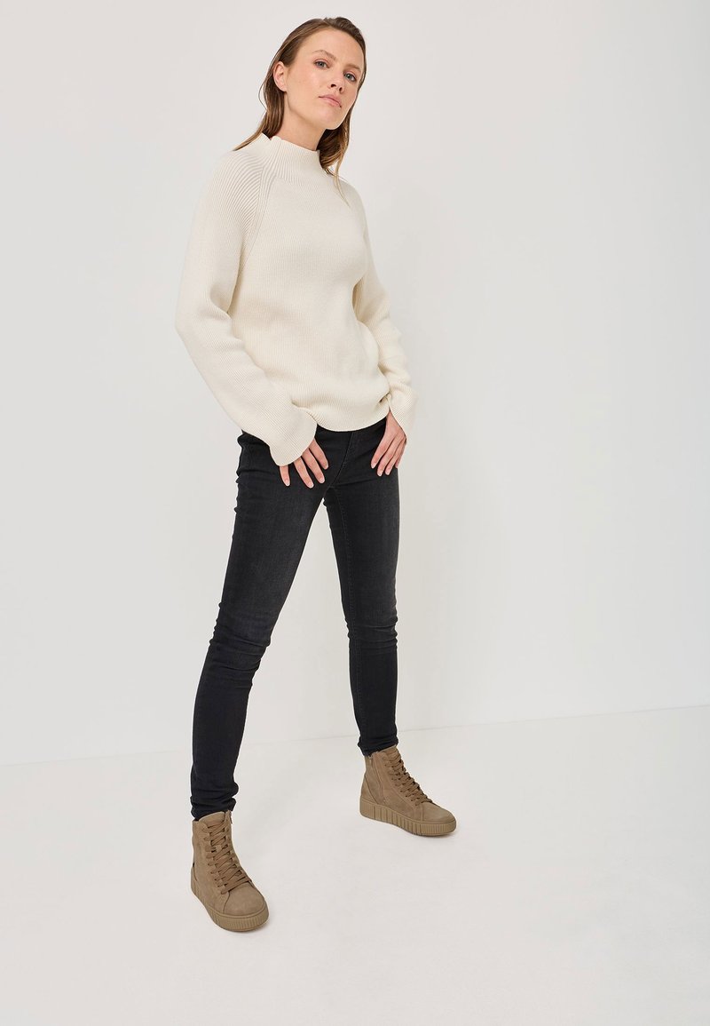 Cremfarbener, gerippter Pullover mit hohem Kragen, kombiniert mit engen schwarzen Jeans und taupefarbenen High-Top-Sneakern. Das Modell steht mit den Händen in den Hüften.