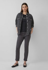 Zwarte denimjack met zilveren knopen, gelaagd over een zwarte top. Grijze broek met opgerolde manchetten en zwarte instappers maken de outfit compleet.