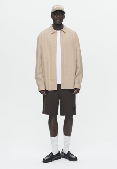Chemise rayée marron clair à boutons, t-shirt blanc, shorts marron, chaussettes blanches, chaussures noires, et une casquette verte avec du texte.