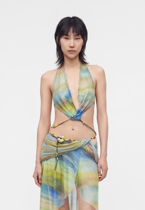 Młoda kobieta nosząca kolorowy, przezroczysty halter top i dopasowaną spódnicę kopertową z koralikowymi zdobieniami, stojąca na jednolitym białym tle.