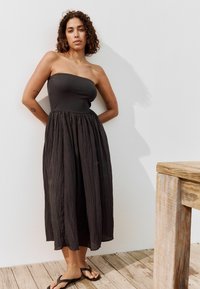 Robe maxi noire sans bretelles avec un corsage ajusté et une jupe en tissu plissé et fluide. Portée avec des sandales noires plates. Mobilier en bois en arrière-plan.