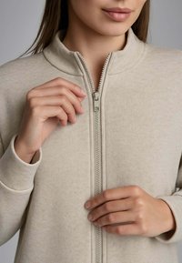 Felpa zip-up beige chiaro con colletto alto, texture morbida e zipper argento. Le mani sono posizionate sulla zip per la chiusura.