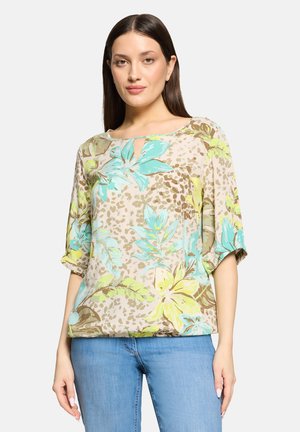 Vrouw met een beige blouse met blauw en geel bloemmotief en lichtblauwe spijkerbroek, staande tegen een effen lichte achtergrond.