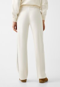 Person trägt lockere weiße Hose mit schwarzem Seitenstreifen und braunen Schuhen, steht vor einem schlichten weißen Hintergrund.