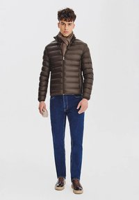 Brązowa ocieplana kurtka z wysokim kołnierzem, zamek błyskawiczny z przodu i prążkowaną fakturą, zestawiona z niebieskimi dżinsami i ciemnymi sneakersami. Szalik zarzucony na szyję.