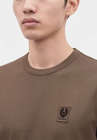 Κοντινό πλάνο ενός ατόμου που φοράει καφέ T-shirt με στρογγυλή λαιμόκοψη και μικρό έμβλημα Belstaff στο στήθος.