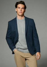Marineblazer mit einem Fischgrätenmuster, getragen über einem hellgrauen Pullover, kombiniert mit beigen Hosen. Glatte Textur, taillierte Passform.
