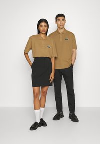 Kaksi henkilöä on pukeutunut yhteensopiviin asuihin, jotka koostuvat ruskeista polo-paitoja logoineen, mustista hameista ja housuista sekä mustista kengistä.