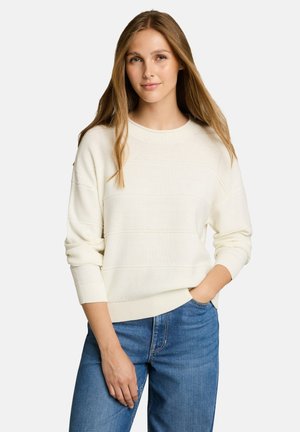 Jonge vrouw met lang lichtbruin haar die een crème gebreide trui en blauwe spijkerbroek draagt, staand met één hand in haar zak tegen een witte achtergrond.