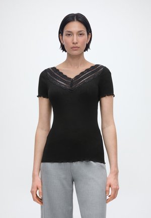 Žena s krátkymi čiernymi vlasmi, oblečená v čiernom krátkoročnom topu s čipkovým výstrihom do V a svetlosivými nohavicami, stojaca pred bielym pozadím.