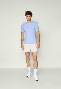 Cămașă sport de culoare albastru deschis, cu un design ajustat, mâneci scurte și accente texturate subtile, purtată cu pantaloni scurți albi și adidași.