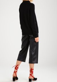 Czarny sweter robiony na drutach, czarne spodnie z sztucznej skóry o kroju 3/4 oraz sandały na obcasie z czerwonymi paskami. Gładka faktura i otwarty krój pleców.