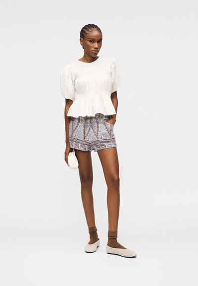 Femme aux cheveux tressés portant un chemisier blanc à manches bouffantes, un short à motifs, des chaussettes marron, des ballerines beige et tenant un petit sac blanc.