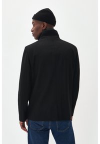 Svart ullblandad kappa med hög krage, strukturerade axlar och rak passform; kombinerad med blå denimjeans och en svart stickad mössa.