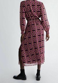 Lång, genomskinlig klänning med ett rosa, lila och guld geometriskt mönster; har en insvängd midja och sidospalt; matchad med svarta skor.