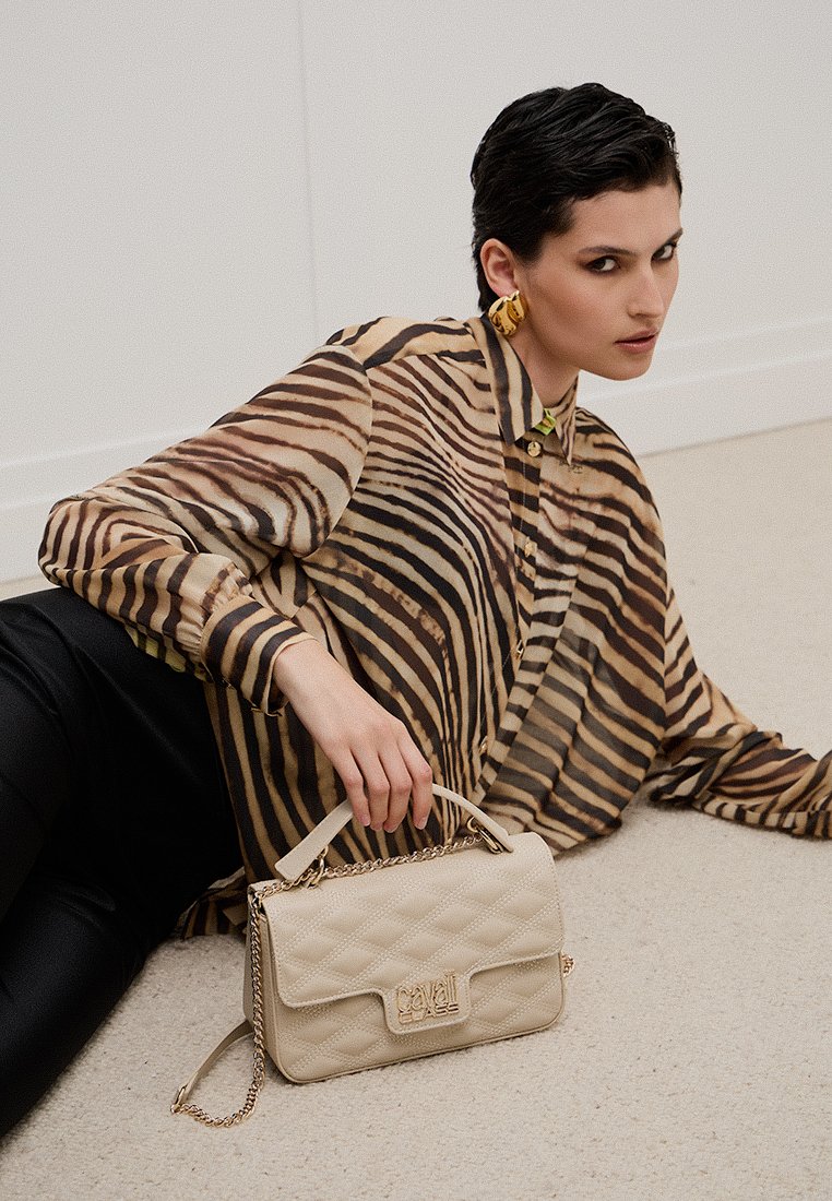 Donna sdraiata sul pavimento che indossa una blusa a strisce tigre e orecchini d'oro, mentre tiene in mano una borsa a tracolla beige con catena e logo dorato.