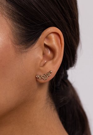 Gros plan sur l'oreille d'une femme aux cheveux foncés attachés, portant une boucle d'oreille dorée texturée de style 'ear crawler' le long du lobe de l'oreille.