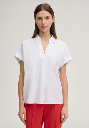 Frau mit glatten braunen Haaren, die eine kurzärmelige weiße Bluse und rote Hose trägt, steht vor einem schlichten hellen Hintergrund.