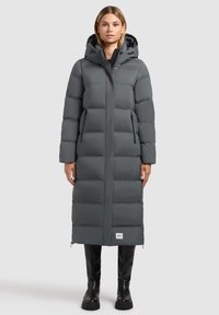 Žena stojící v dlouhém tmavě šedém zatepleném zimním kabátu s kapucí, na zip, a černých kozačkách nad kolena.