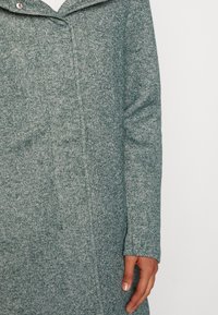 Cappotto verde in tweed con collare a scollo a V, chiusura frontale con bottoni e tessuto testurizzato. Presenta maniche lunghe e dettagli di pattern discreti.
