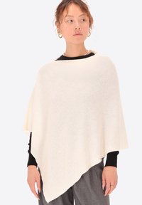 Poncho color crema chiaro realizzato in tessuto morbido e testurizzato, con forma triangolare e scollatura leggermente arrotondata, indossato sopra una maglietta nera.
