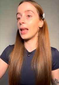 Frau mit langen, glatten Haaren, die auf einer Seite befestigt sind, trägt ein dunkles Oberteil und spricht vor einem neutralen Hintergrund.
