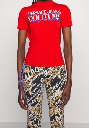 Rotes kurzärmliges T-Shirt mit dem Aufdruck "VERSACE JEANS COUTURE" in fettem Blau und Weiß, kombiniert mit schwarzen Hosen, die mit goldenen und weißen Textmustern verziert sind.