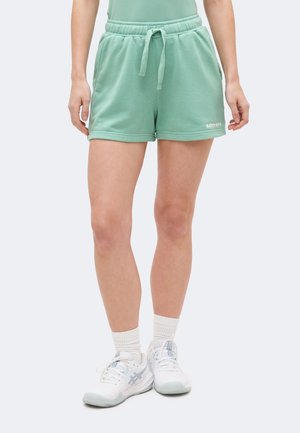 Γυναίκα που φοράει σορτς μέντας με κορδόνια, λευκές κάλτσες μέχρι τον αστράγαλο και λευκά αθλητικά παπούτσια, στέκεται μπροστά σε απλό φόντο.