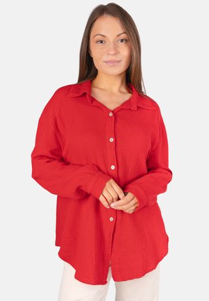 Frau mit glattem braunem Haar, die ein lockeres rotes Hemd mit Knöpfen und weiße Hosen trägt, steht mit sanft verschränkten Händen.
