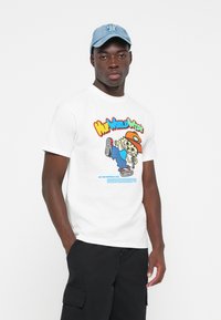 White cotton t-shirt featuring a colorful graphic design with cartoon character and text. Model wears black cargo shorts and a light blue cap.