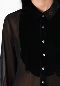 Camisa preta com colarinho e painel frontal em veludo, mangas transparentes e fecho de botão com botões em tom prateado. Detalhe de tecido texturizado.