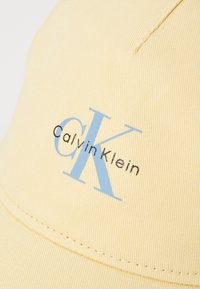 Casquette jaune en tissu texturé, ornée d'un logo "CK" bleu et de "Calvin Klein" en noir. La couture le long de la visière est visible.