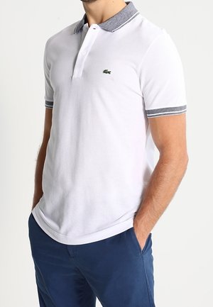 Homme portant un polo blanc avec col et bords des manches gris, un pantalon bleu, les mains dans les poches.