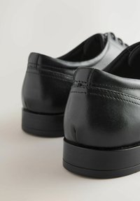 Lakkitud mustad nahast dressiketsid ühtsetel kontsadelt, vaadatuna tagantpoolt heledal taustal.