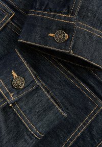 Tumedat sinist teksajakki lähivaade, mille varrukatel on beežid õmblused ja messingist nööbid, millele on pressitud "JJXX" ja tähed.