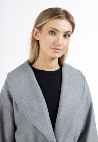 Texturovaný šedý kabát s širokými klopami, vyrobený z měkkého materiálu. Nositelný přes černý top, který má uvolněný střih.