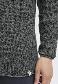 Grau und schwarz gestrickter Pullover mit einem strukturierten Muster, einem umgeschlagenen Bündchen und einem kleinen Etikett am Saum. Das Material wirkt weich und warm.