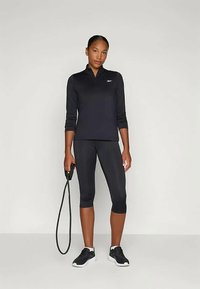 Camisola atlética preta de mangas longas com gola de fecho, combinada com leggings capri pretas. Ténis pretos e uma trela de cachorro na mão.