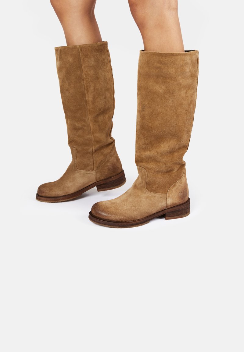 Bottes en suède beige jusqu'au genou, avec un orteil rond et un talon empilé. Présentent une texture lisse et des détails de couture subtils sur les côtés.