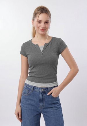Νέα γυναίκα με ξανθά μαλλιά σε κοτσίδα, φορά γκρι μπλουζάκι τύπου henley με κοντά μανίκια πάνω από ανοιχτό γκρι τοπ και μπλε τζιν, ποζάροντας με το ένα χέρι στην τσέπη.