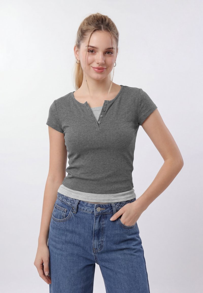Νέα γυναίκα με ξανθά μαλλιά σε κοτσίδα, φορά γκρι μπλουζάκι τύπου henley με κοντά μανίκια πάνω από ανοιχτό γκρι τοπ και μπλε τζιν, ποζάροντας με το ένα χέρι στην τσέπη.