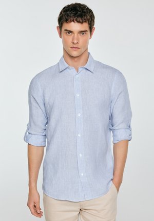 Młody mężczyzna o kręconych brązowych włosach, ubrany w jasnoniebieską koszulę z podwiniętymi rękawami i beżowe spodnie, stojący na jednolitym tle.