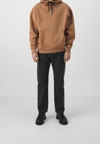 Sudadera marrón con cordón y bolsillo frontal, combinada con jeans negros de pierna recta y zapatos negros con cordones.