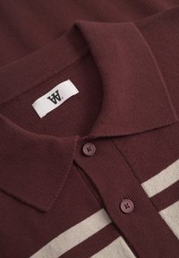 Nærbillede af en bordeaux strikket polotrøje-krave med to knapper og et hvidt mærke, der har to horisontale cremefarvede striber på stoffet.